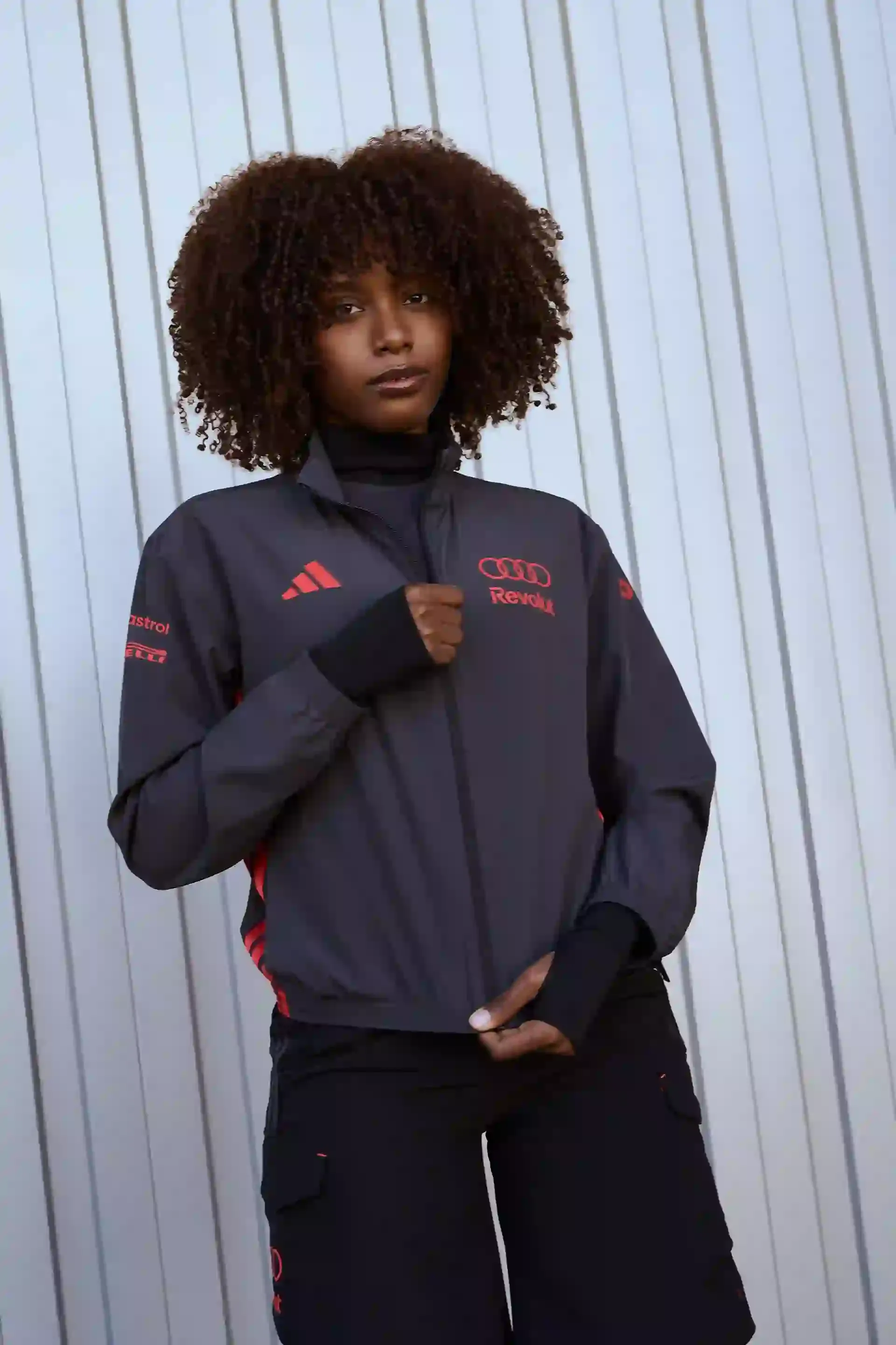 Portrait of a model with curly hair dark team jacket, with lava red adidas Logo and Audi rings Logo, neutral background confident posture modern performance look