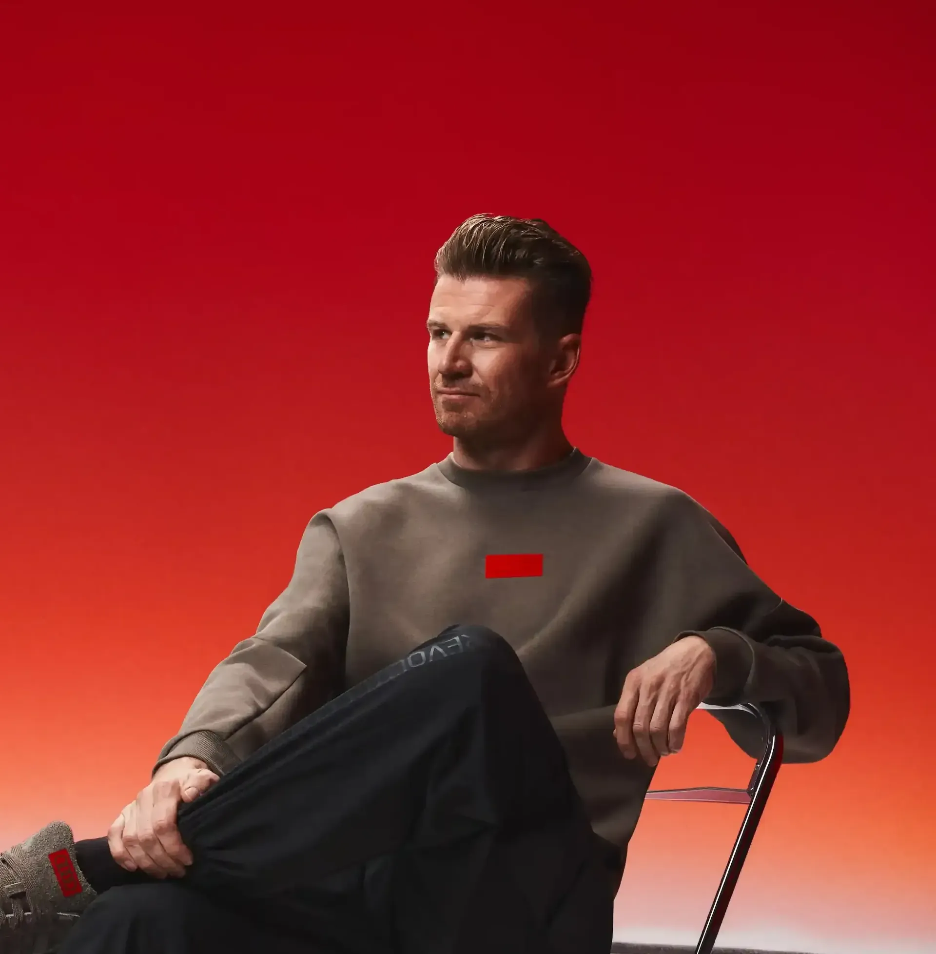 Formula 1 athlete Nico Hülkenberg sitting on a chair, relaxed pose, black pants, brown long-sleeve shirt, minimalist studio setup with motorsport graphics on the floor.