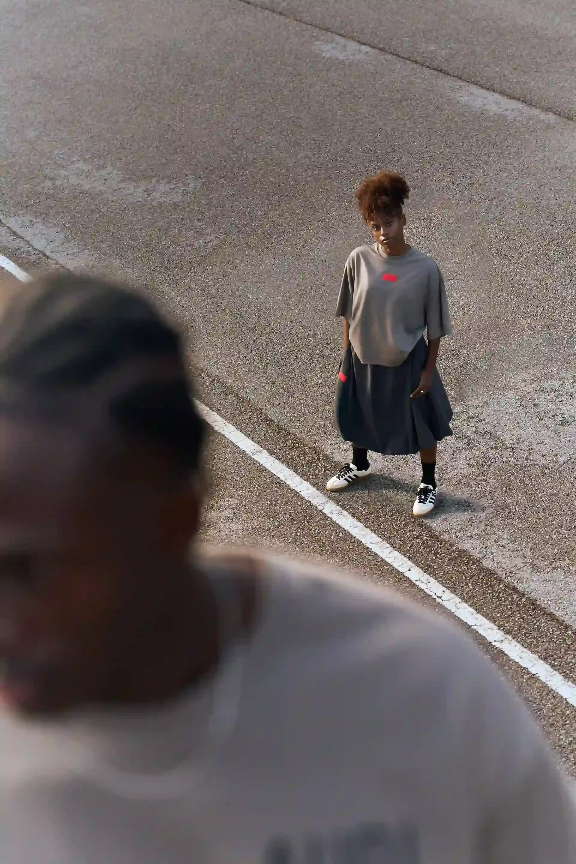Two models, the one in front shown in closeup, standing on a concrete road, wearing gray-brown shirts and a dark skirt, relaxed posture with a modern streetwear look.
