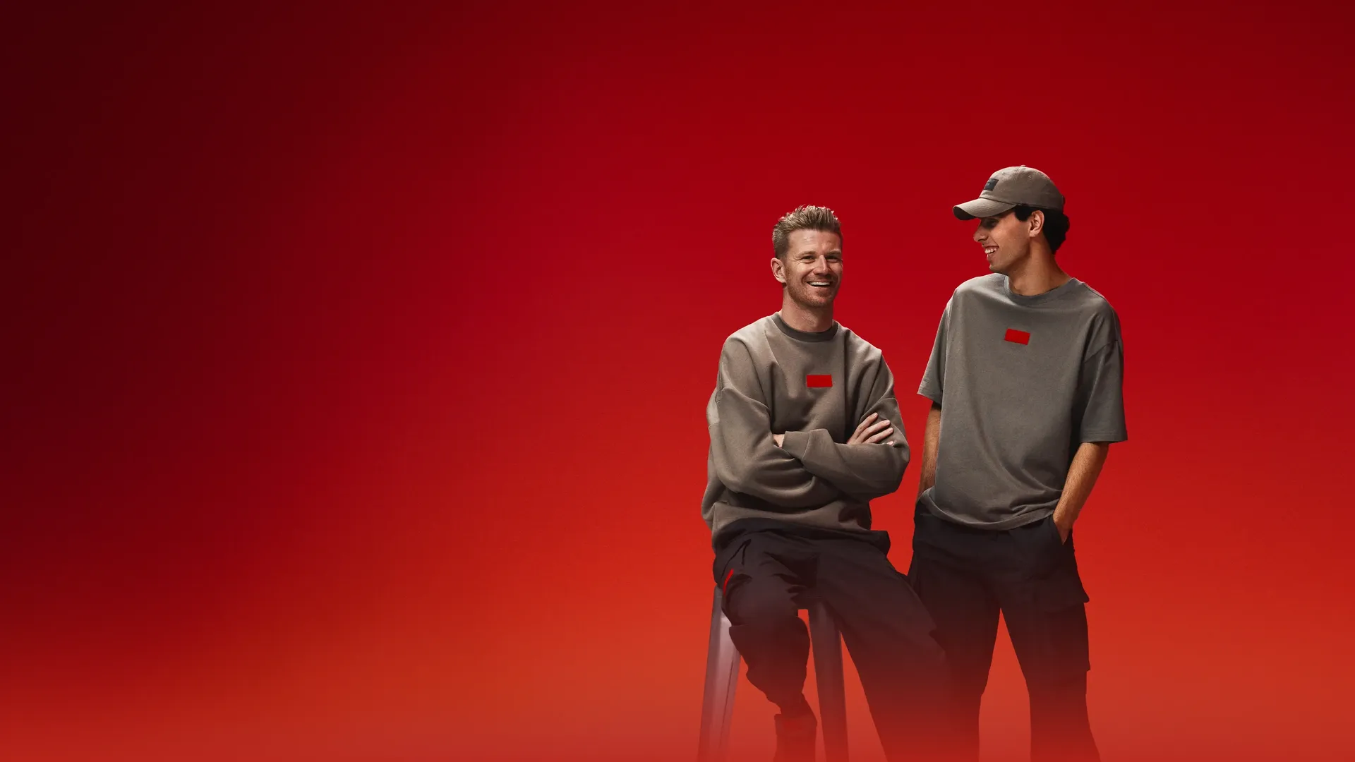 Two F1 Drivers, Nico Hulkenberg and Gabriel Bortoleto, in coordinated taupe, streetwear, outfits pose in a studio with a red gradient background; one sits on a stool smiling while the other stands beside him, looking over.
