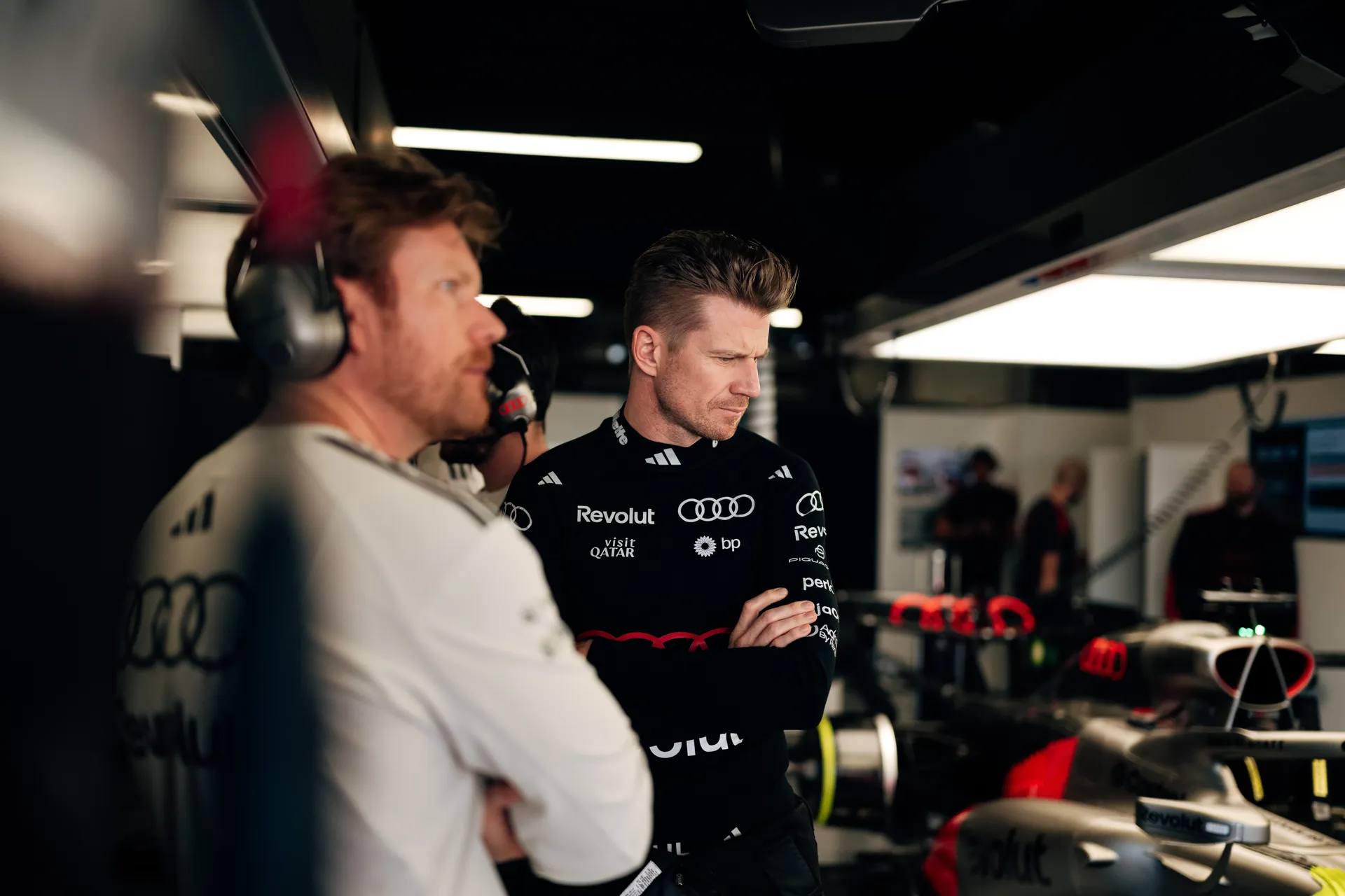 Nico Hulkenberg standing in the Audi Revolut F1® Team garage next to engineers, observing the car with arms crossed.