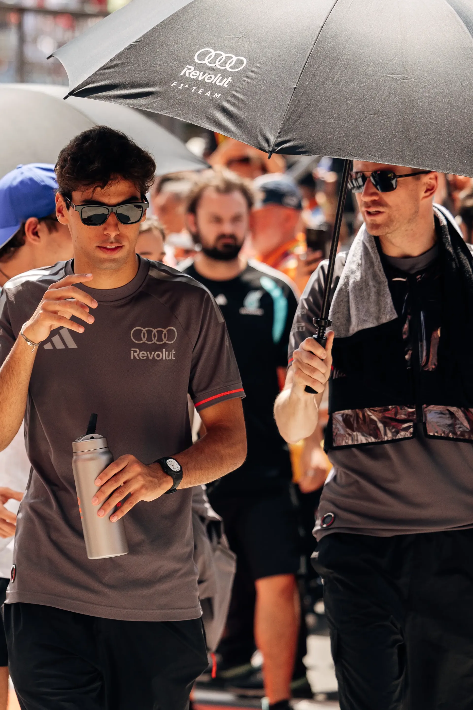 Gabriel Bortoleto walks through the paddock during the Hungarian Grand Prix drivers' parade carrying a drink, alongside Nico Hulkenberg under an Audi umbrella, both wearing Audi Revolut F1® Team shirts.