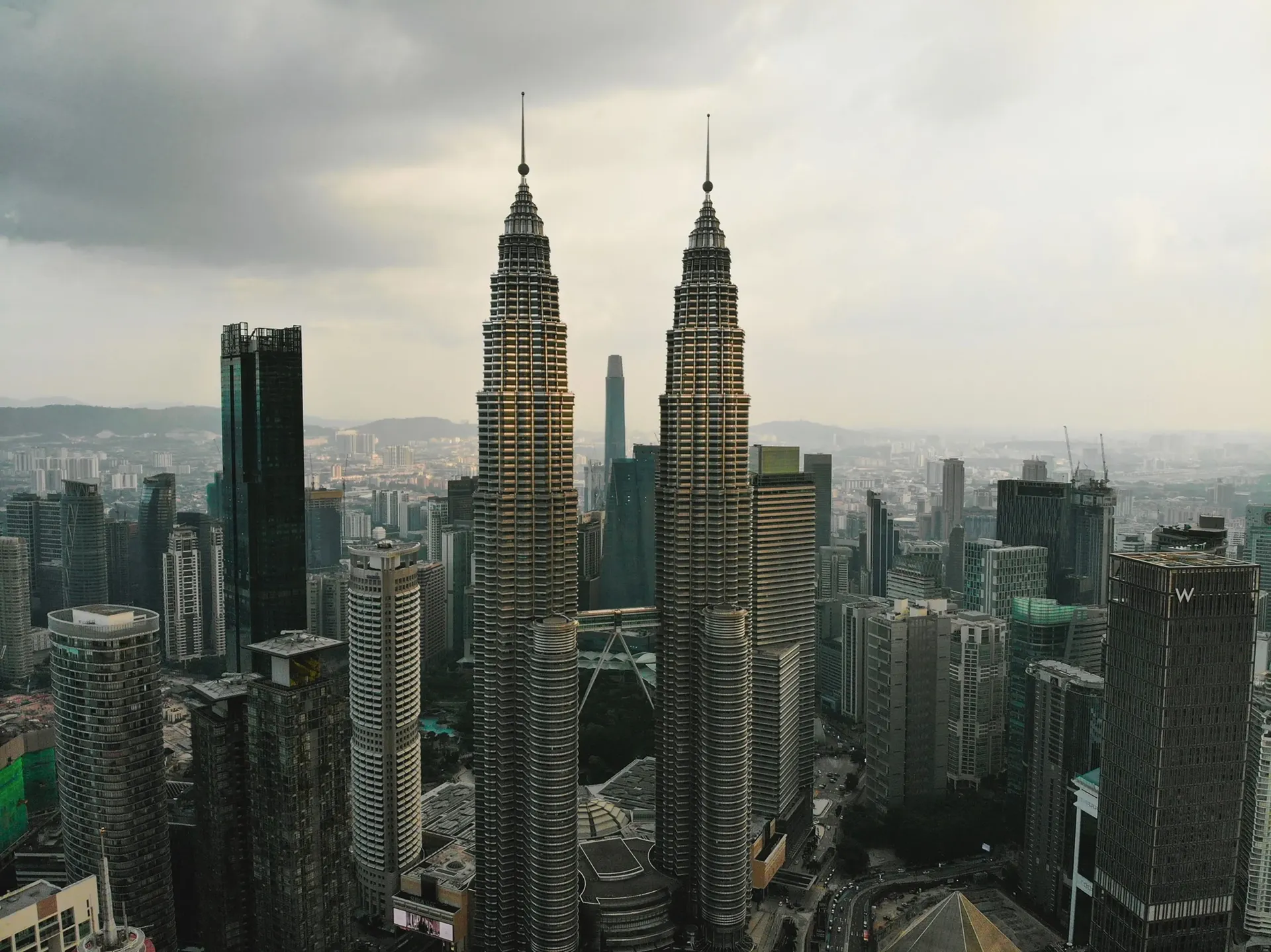 A drone view of the Petronas Twin Towers rising above the Kuala Lumpur skyline, surrounded by modern skyscrapers, lush green spaces, and the city's urban landscape in Malaysia.