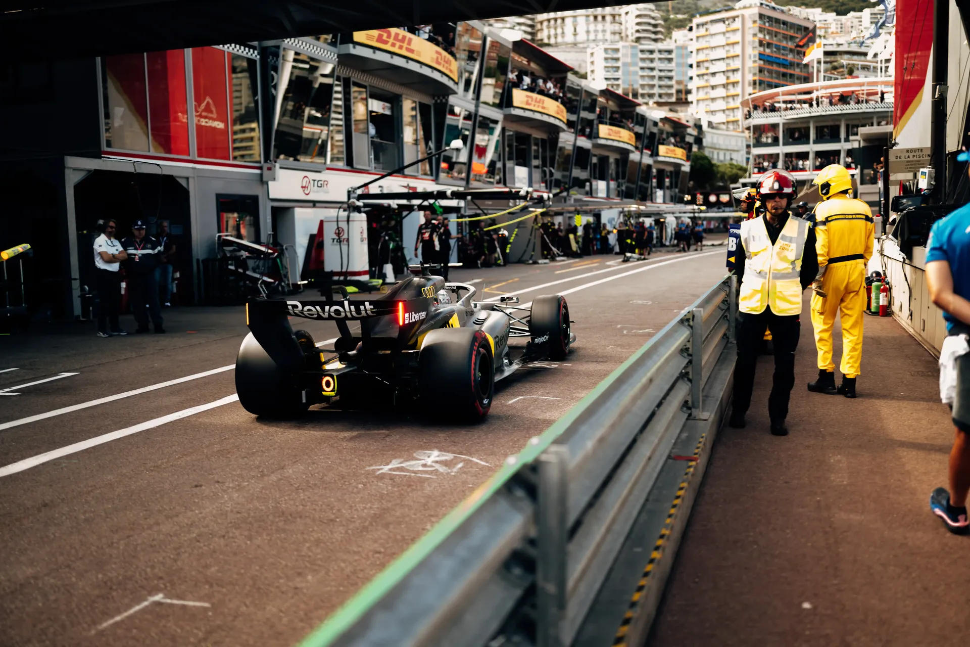 Audi Revolut F1® Team car R26 driving down the Monaco pit lane with marshals and team garages alongside.