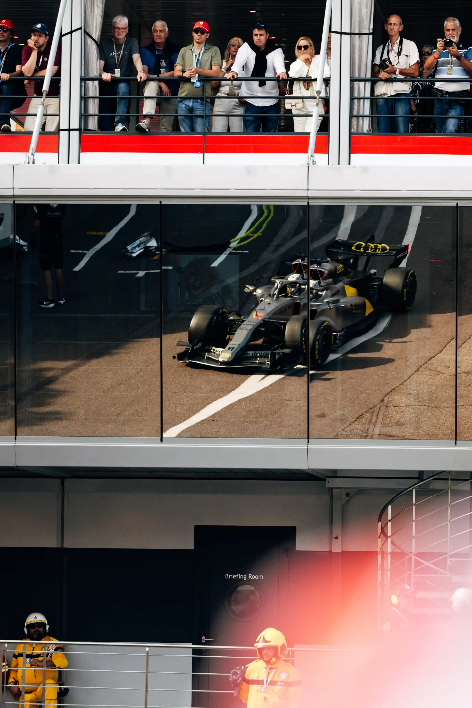 Audi Revolut F1® Team car R26 reflected in the glass facade of the Monaco pit building with spectators above.