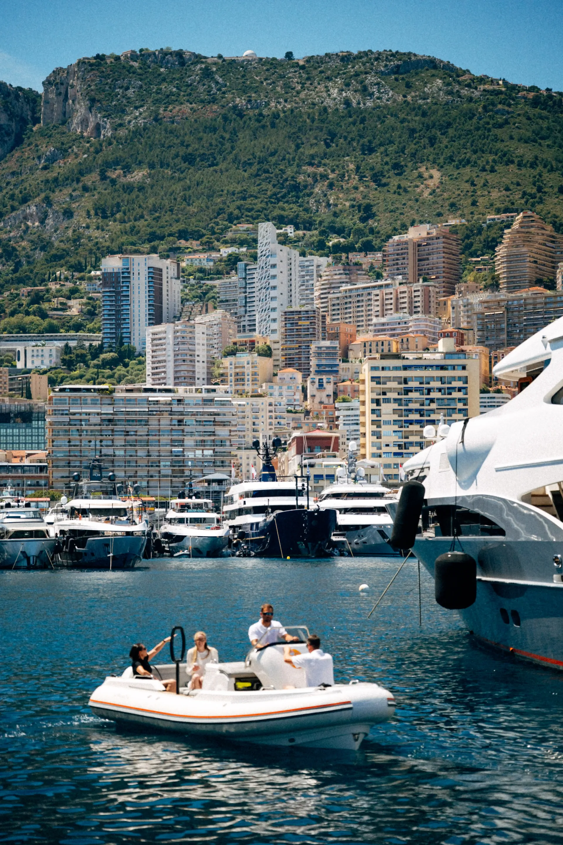 Luxury yachts anchored in Monaco harbor with green hills and waterfront residences behind.