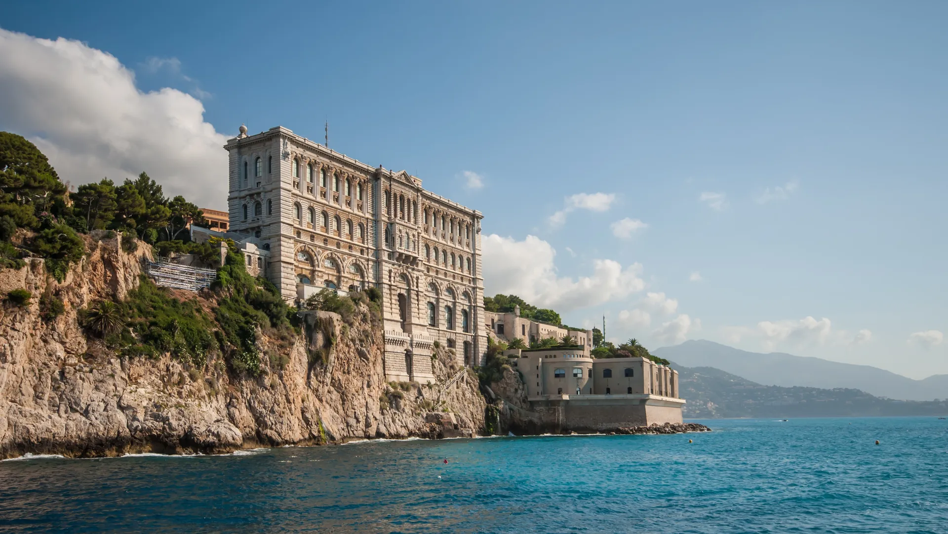 Historic waterfront building in Monaco overlooking the Mediterranean Sea, with mountains in the distance.